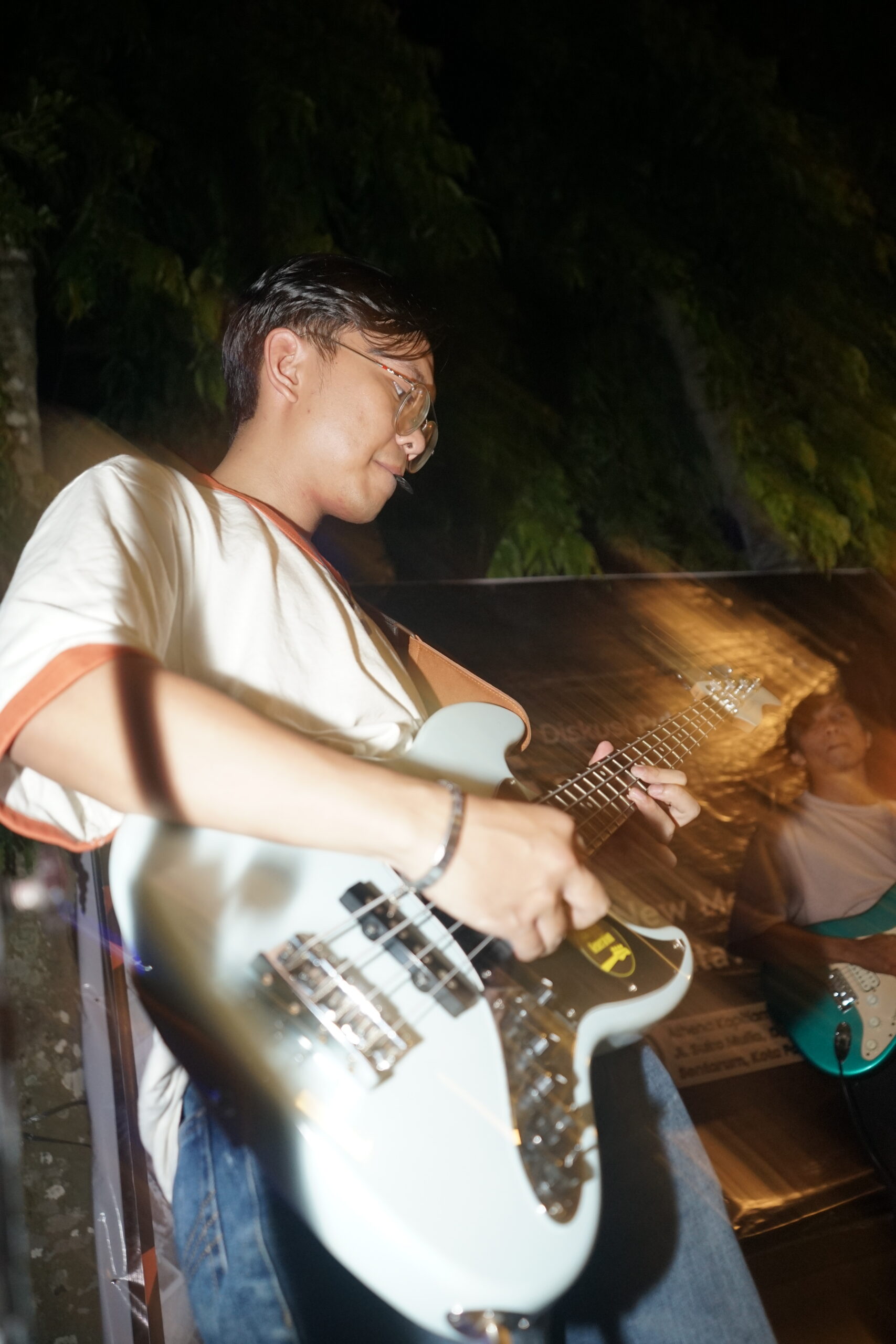 Foto dari sudut bawah (low angle) seorang basis berkacamata yang sedang fokus memainkan gitar bass elektrik berwarna putih abu-abu pada sesi musik akustik malam hari. Di latar belakang yang sedikit buram, tampak seorang gitaris lain sedang memainkan gitar elektrik berwarna biru toska di area panggung luar ruangan.