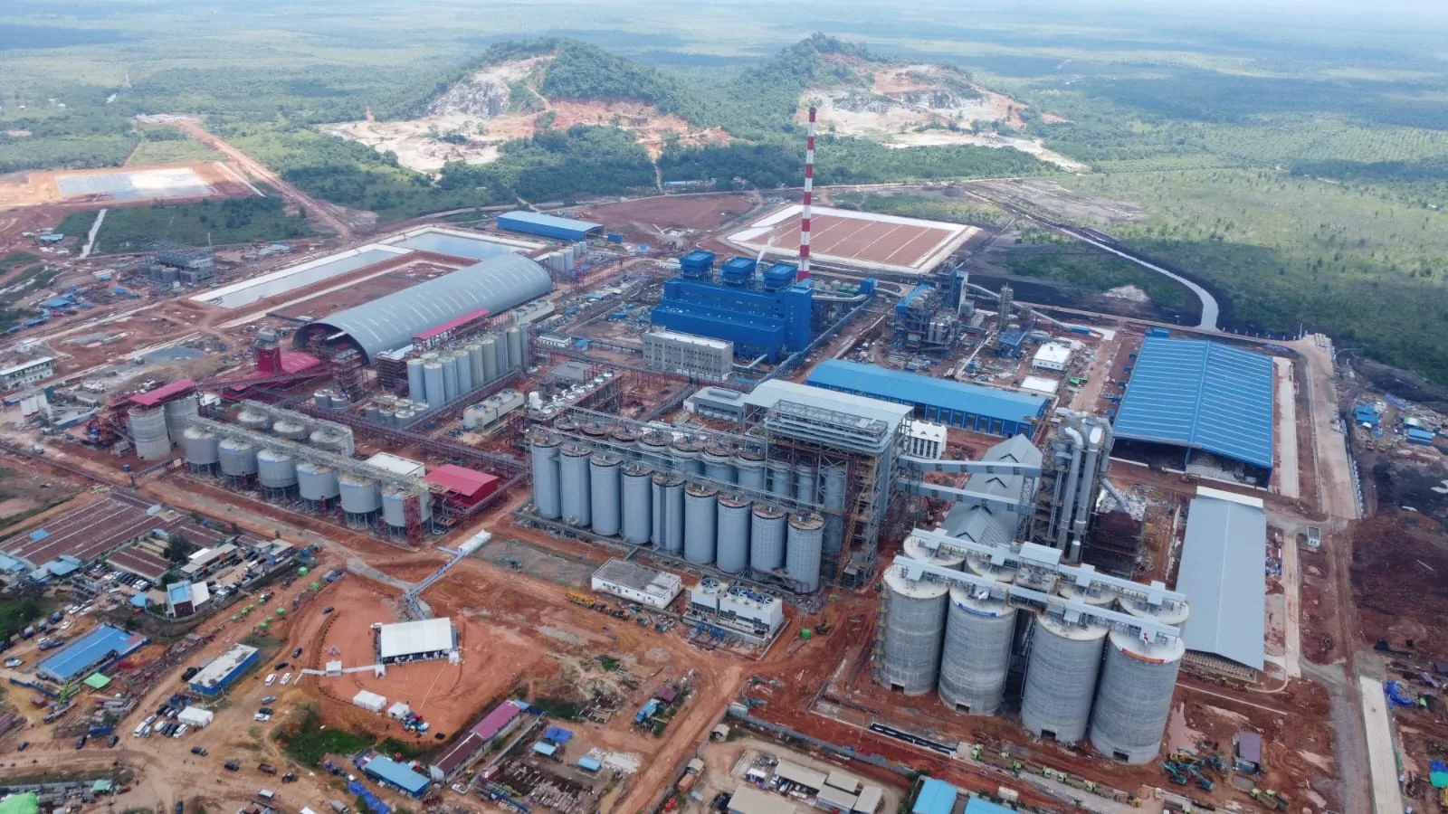 Pemandangan udara (aerial view) sebuah kompleks pabrik industri besar dengan tangki-tangki penyimpanan perak, gedung-gedung biru, dan cerobong asap tinggi di tengah kawasan hijau.