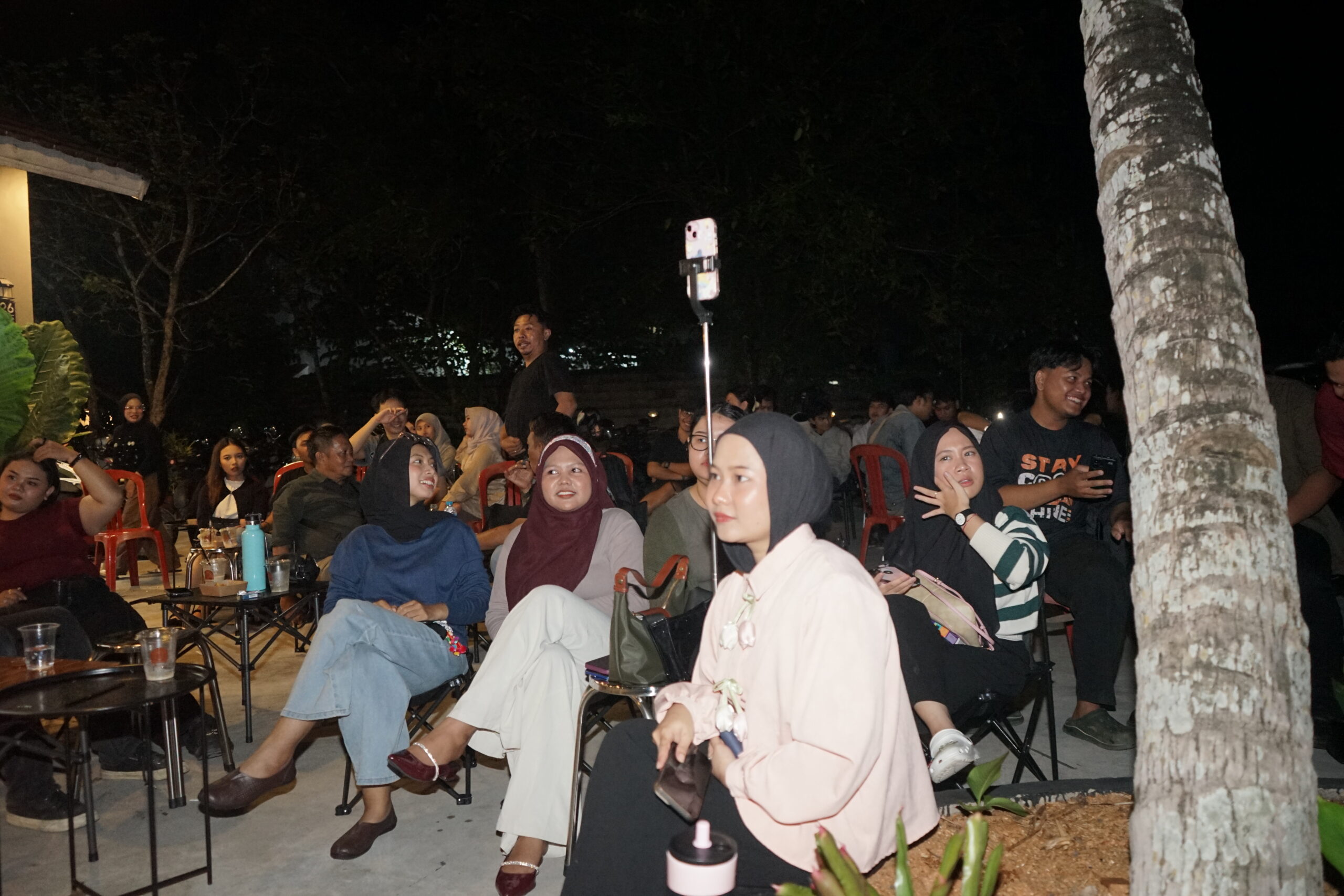 Foto suasana malam sekumpulan audiens muda, termasuk beberapa wanita berhijab di baris depan, yang sedang duduk di kursi lipat outdoor sambil menyimak jalannya diskusi publik di sebuah cafe di Pontianak