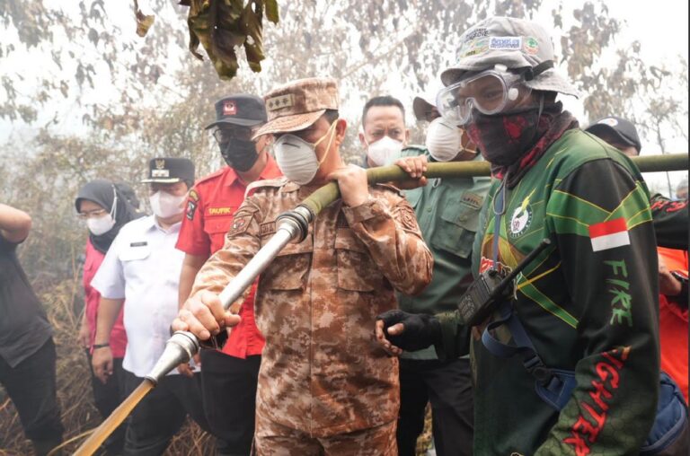 Gubernur Kalimantan Barat mengenakan seragam loreng dan masker berdiri di tengah kepulan asap sambil memegang selang air bersama seorang petugas pemadam kebakaran berseragam hijau, dikelilingi sejumlah pejabat dan petugas lainnya di lokasi karhutla.