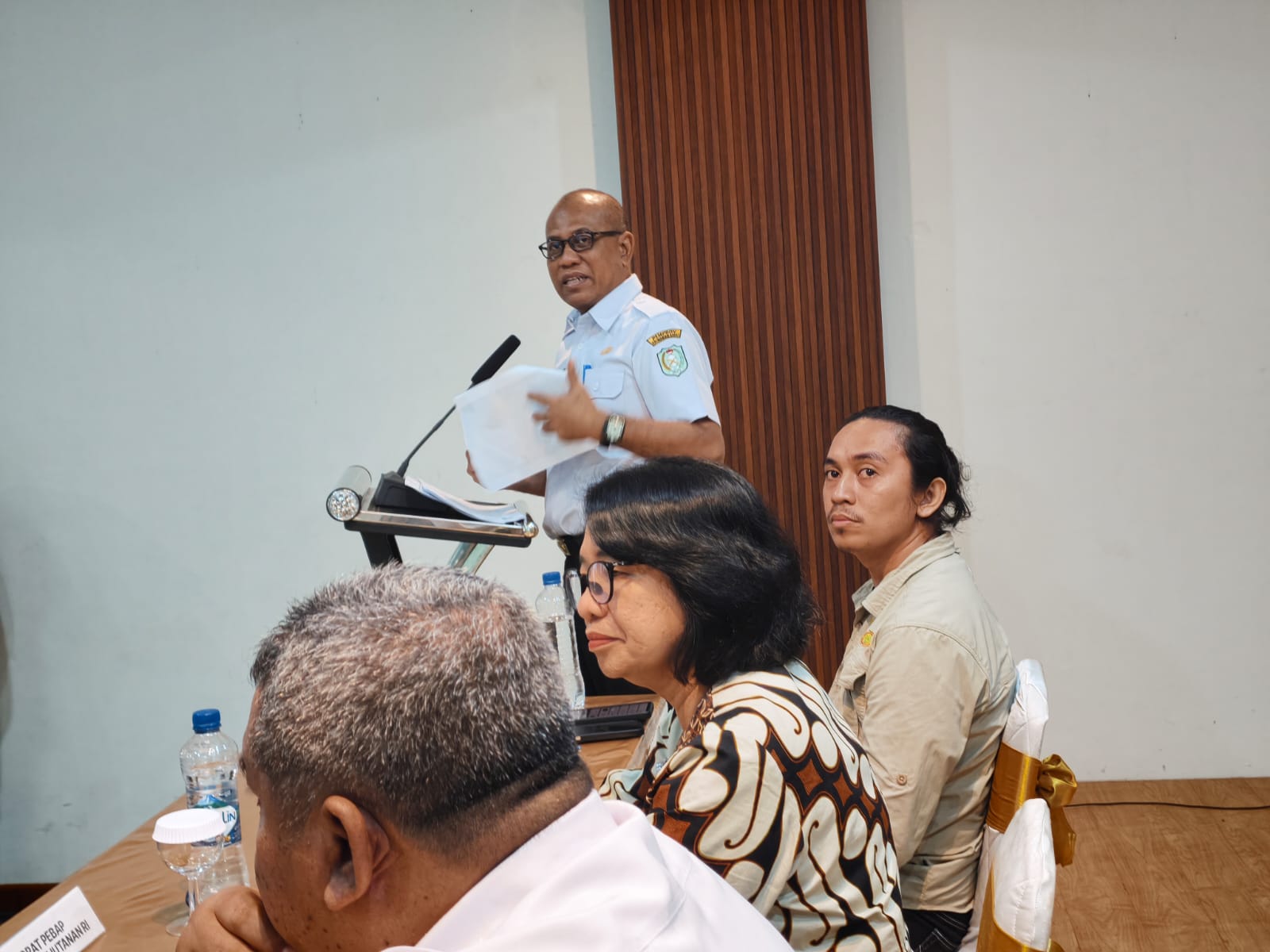 Seorang pejabat berseragam dinas putih dan berkacamata berbicara di podium sambil memegang kertas, dengan peserta duduk di meja narasumber di depannya