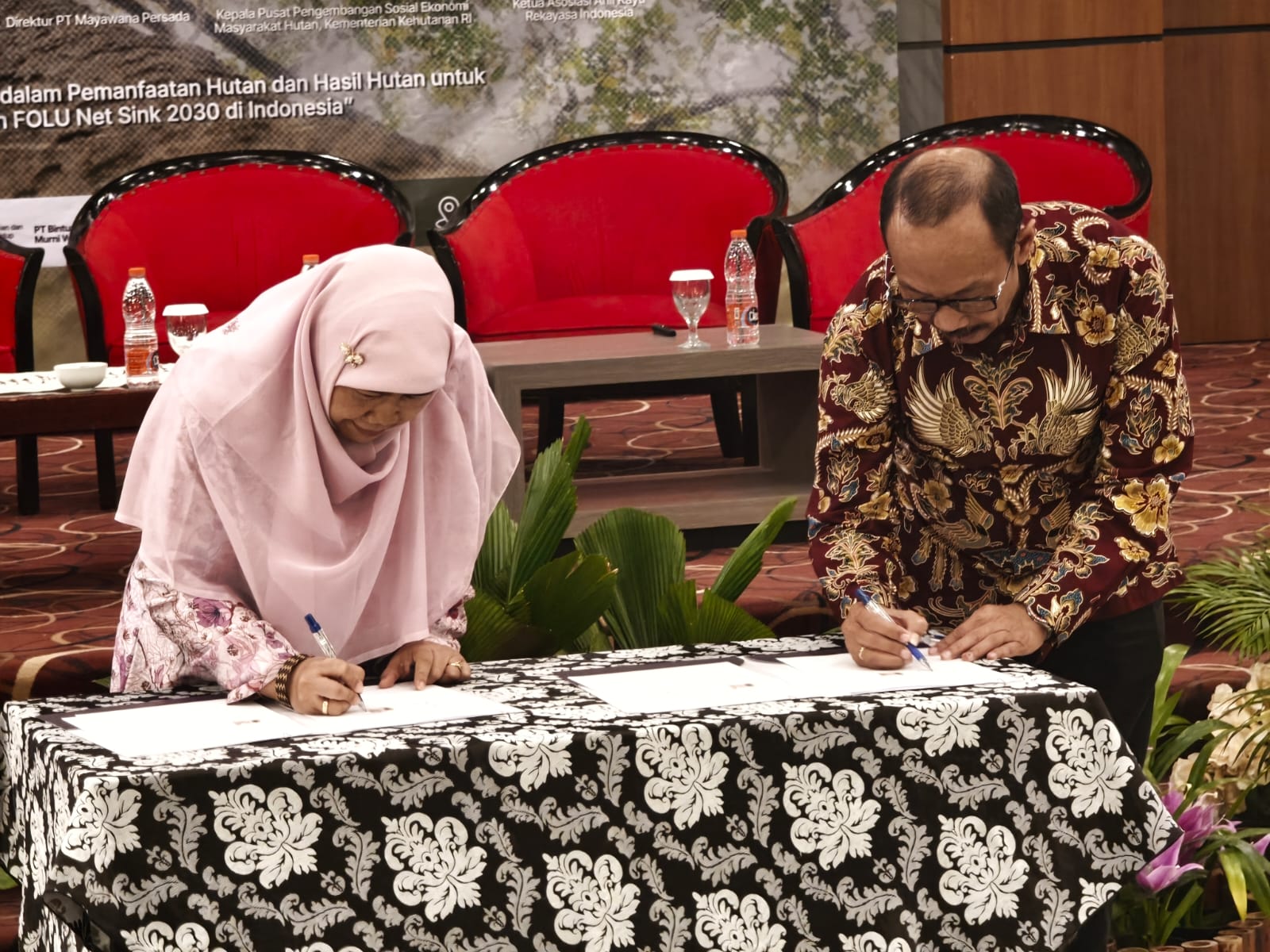 Seorang perempuan berhijab merah muda dan seorang pria berbatik menandatangani dokumen kerja sama di atas meja berkain motif.