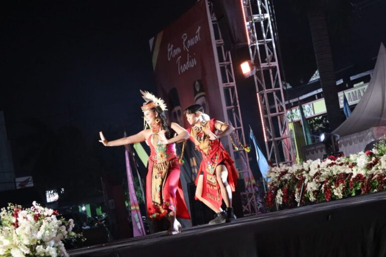Penampil berbusana adat dan berhiasan bulu menari dan berlutut di halaman pada malam hari saat pentas seni budaya.