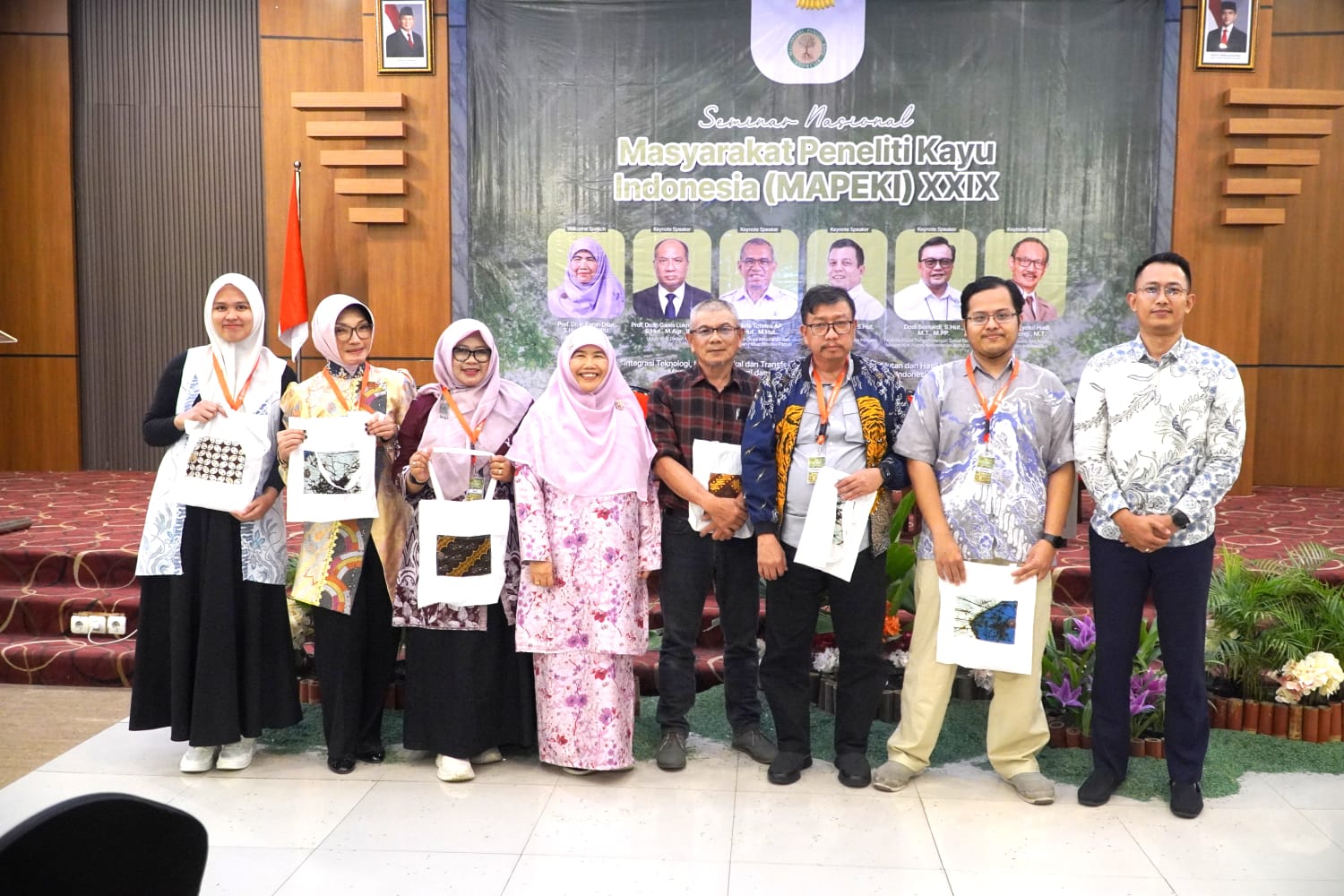 Delapan peserta dari Untan berdiri berjajar membawa tas kain batik di depan latar Seminar Nasional MAPEKI XXIX.