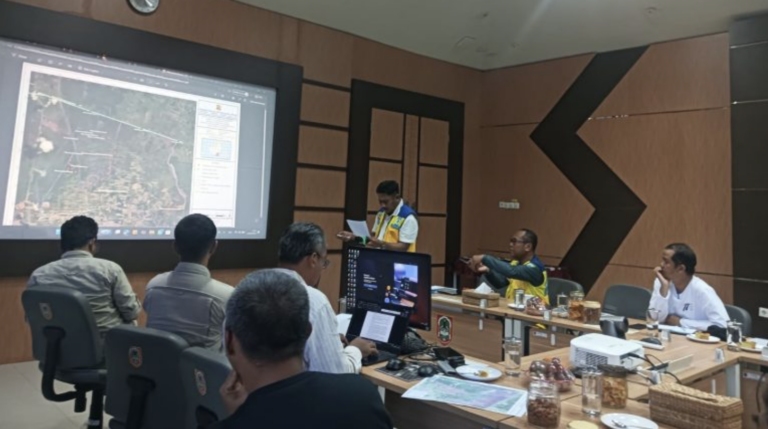 Peserta rapat menyimak paparan peta citra satelit di layar besar, dengan seorang petugas berompi berdiri membaca catatan.
