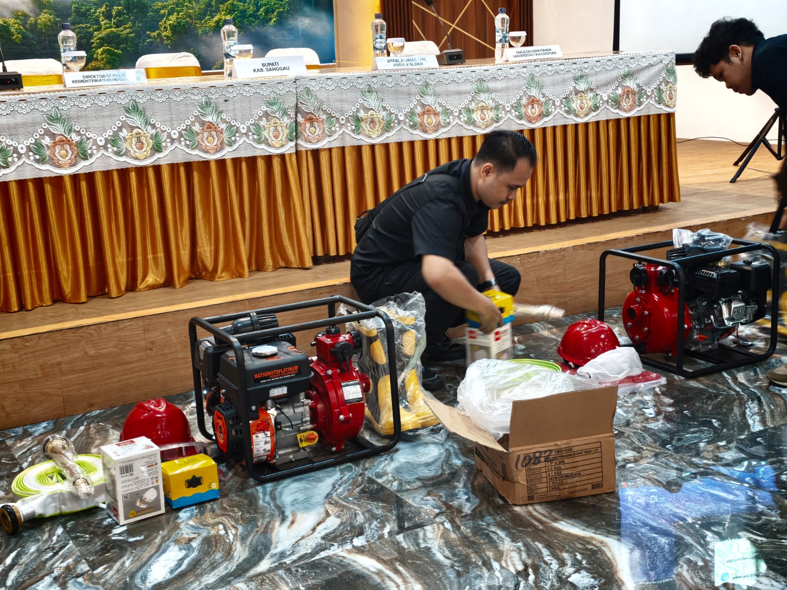 Seorang petugas berkemeja hitam menata peralatan pemadam kebakaran berupa dua pompa air merah, selang, helm merah, dan kardus di depan meja narasumber