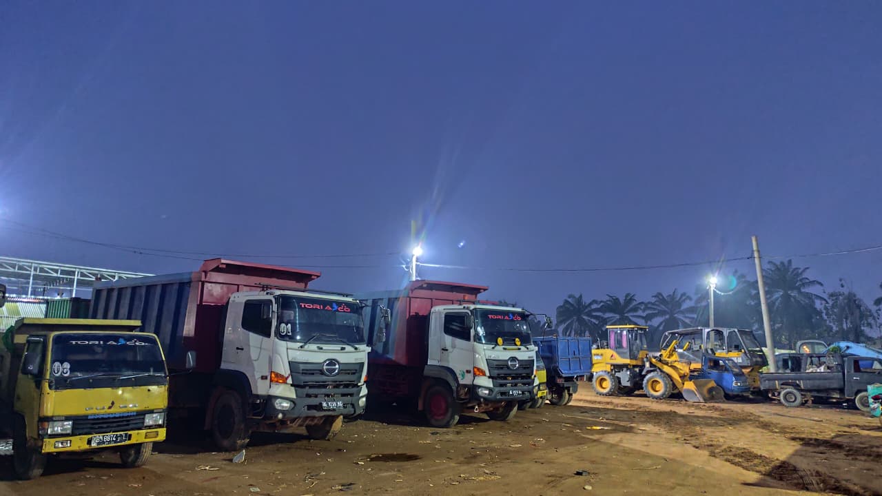 Deretan truk bak bertulisan Tori Asco dan sebuah wheel loader kuning terparkir di pangkalan pada malam hari.